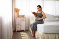 Happy Asia pregnant woman reading book and sitting on white bed Royalty Free Stock Photo