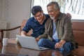 happy asian adult son and senior father sitting on sofa using laptop together at home . young man teaching old dad using internet Royalty Free Stock Photo