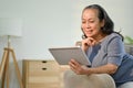 Happy Asian-aged retired woman watching movie on her digital tablet on sofa Royalty Free Stock Photo