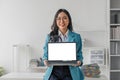 Happy asian business woman holding laptop with blank screen stands in a modern office looking at the camera and smiling Royalty Free Stock Photo