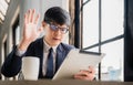 Happy asian businessman in suit wearing glasses talk and waving while video conference call from digital tablet in coffee shop