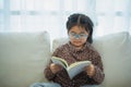 Happy asian child baby girl smiling wearing glasses and reading book while sitting on couch sofa in living room at home. Girl Royalty Free Stock Photo