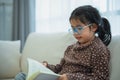 Happy asian child baby girl smiling wearing glasses and reading book while sitting on couch sofa in living room at home. Girl Royalty Free Stock Photo