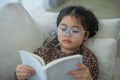 Happy asian child baby girl smiling wearing glasses and reading book while sitting on couch sofa in living room at home. Girl Royalty Free Stock Photo