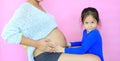 Happy asian child girl with belly of her mom with love. Little girl relaxing with pregnant mother isolated on pink background.