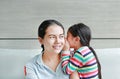 Happy Asian child girl sharing a secret to her mom in the living room at home. Kid girl whispering gossip a something to mother