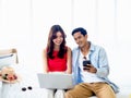 Happy Asian couple, young man and woman in casual looking at smart mobile phone screen while using and working with laptop. Royalty Free Stock Photo