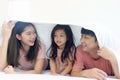 Happy Asian family has fun in bedroom. Father, mother daughter lying in bed together and hiding under white blanket. Parents and Royalty Free Stock Photo