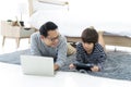 Happy Asian father using a laptop computer and his cute little son using a digital tablet while lying on the carpet in bedroom. Royalty Free Stock Photo