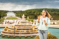 Asian girl snacking on a croissant and drinking coffee in front of the Palace and Park complex of Versailles, in France