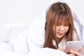 Happy Asian girl using a mobile phone lying under a blanket on the bed at home. Social and technology concept Royalty Free Stock Photo