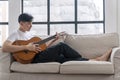Happy asian handsome man playing acoustic guitar sitting on sofa relax beside window at apartment. Young male enjoying play string Royalty Free Stock Photo