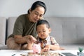 Happy Asian kid and mom saving money together, putting cash into piggy bank. Mother playing with child on heating floor at home, Royalty Free Stock Photo