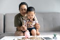 Happy Asian kid and mom saving money together, putting cash into piggy bank. Mother playing with child on heating floor at home, Royalty Free Stock Photo