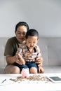 Happy Asian kid and mom saving money together, putting cash into piggy bank. Mother playing with child on heating floor at home, Royalty Free Stock Photo