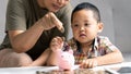 Happy Asian kid and mom saving money together, putting cash into piggy bank. Mother playing with child on heating floor at home, Royalty Free Stock Photo