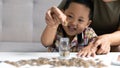 Happy Asian kid and mom saving money together, putting cash into piggy bank. Mother playing with child on heating floor at home, Royalty Free Stock Photo