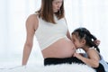 Happy Asian little cute kid girl playing with pregnant mom on bed at home, touch and kiss mother belly with tender and love. Royalty Free Stock Photo