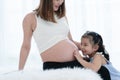 Happy Asian little cute kid girl playing with pregnant mom on bed at home, touch mother belly and navel with tender and love. Royalty Free Stock Photo