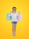 Happy Asian little girl wearing sunglasses holding shopping bags on yellow isolated background with clipping path. Full length Royalty Free Stock Photo