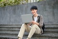 Happy Asian male college student working on his school project, using laptop on campus outside stairs Royalty Free Stock Photo