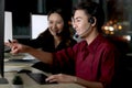 Happy Asian man with headphones work night shift at call center customer care service desk with colleagues, point at computer Royalty Free Stock Photo