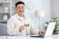 Happy Asian man sitting in bright office at table in front of laptop, holding card in hand, satisfied with online Royalty Free Stock Photo