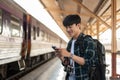 A happy Asian man tourist using his smartphone while standing at a platform in a railway station Royalty Free Stock Photo