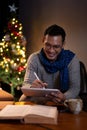 A happy Asian man in a warm cosy clothes is using his tablet while sitting beside a Christmas tree