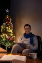 A happy Asian man in a warm cosy clothes is using his tablet while sitting beside a Christmas tree