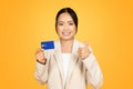 Happy Asian millennial lady in suit, holds credit card make thumb up gesture