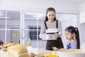 Happy Asian mother teaching her young daughter to bread baking in white modern kitchen while sieving wheat flour for mixing