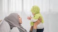 Happy asian muslim mother holding adorable little baby daughter wearing hijab in her arms on white bed in bedroom Royalty Free Stock Photo