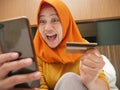 Happy Asian muslim woman wearing hijab smiling and laughing, when making purchase on online shop with her phone and credit card Royalty Free Stock Photo