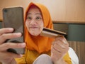 Happy Asian muslim woman wearing hijab smiling and laughing, when making purchase on online shop with her phone and credit card Royalty Free Stock Photo