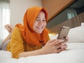 Happy Asian muslim woman wearing hijab smiling when reading text message or chat on her phone while lying on bed Royalty Free Stock Photo