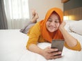 Happy Asian muslim woman wearing hijab smiling when reading text message or chat on her phone while lying on bed Royalty Free Stock Photo