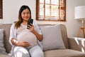 Happy Asian pregnant woman using her phone while relaxing on sofa in her living room Royalty Free Stock Photo