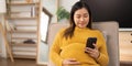 Happy Asian pregnant woman using her phone while relaxing on sofa in her living room Royalty Free Stock Photo