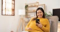 Happy Asian pregnant woman using her phone while relaxing on sofa in her living room Royalty Free Stock Photo