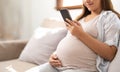 Happy Asian pregnant woman using her phone while relaxing on sofa in her living room Royalty Free Stock Photo