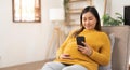 Happy Asian pregnant woman using her phone while relaxing on sofa in her living room Royalty Free Stock Photo