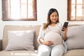 Happy Asian pregnant woman using her phone while relaxing on sofa in her living room Royalty Free Stock Photo