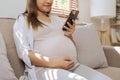 Happy Asian pregnant woman using smart phone while relaxing on sofa in her living room Royalty Free Stock Photo