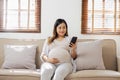 Happy Asian pregnant woman using smart phone while relaxing on sofa in her living room Royalty Free Stock Photo