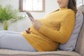 Happy Asian pregnant woman using smart phone while relaxing on sofa in her living room Royalty Free Stock Photo