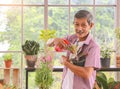 Asian senior man with mustache standing at table indoor with plant pots in his arm and spray water to it, smiling and looking at Royalty Free Stock Photo