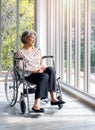 Happy Asian senior woman in casual dress sits in wheelchair, smile and looking out of the glass window with natural green view. Royalty Free Stock Photo