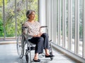 Happy Asian senior woman in casual dress sits in wheelchair, smile and looking out of the glass window. Royalty Free Stock Photo