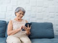 Happy Asian senior woman sit on couch, holding and looking at digital tablet screen in hand in living room. Royalty Free Stock Photo